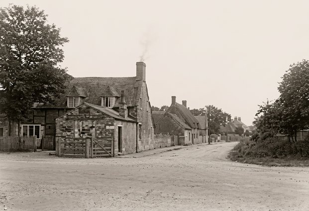 Old sepia postcard of the village street in Northborough, with stone cottages and trees either side of the road.