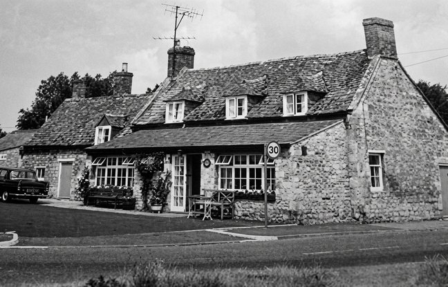 Black and white photograph of The Pack Horse: a long stone building with three dormer windows in the roof, bay windows and an old car parked outside.